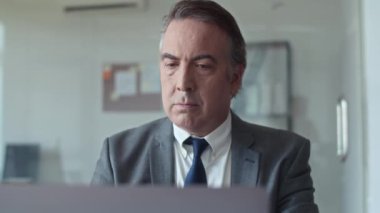 Dizüstü bilgisayarda çalışan ve ofisteki kameraya gülümseyen yaşlı finans müdürünün orta boy portresi.