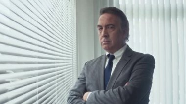 Pencerenin yanında duran ve kameraya bakan üst düzey ofis çalışanının orta boy portresi.