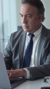 Ofisteki dizüstü bilgisayarda çalışan, kameraya bakan olgun iş adamının dikey portresi.