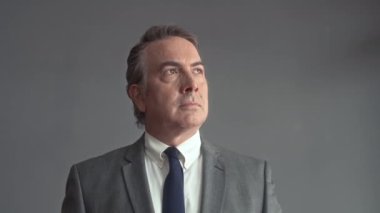 Gri duvar arkasında duran kameraya bakan neşeli kıdemli işadamının orta boy yakın portresi.