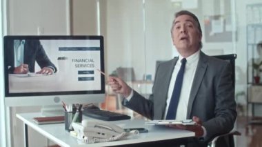 Orta yaşlı girişimcinin finansal hizmetlerin iniş sayfasıyla bilgisayar ekranını göstermesi ve kamerada konuşması.