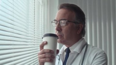 Beyaz önlüklü ve gözlüklü yaşlı bir doktor hastanede ameliyattan sonra dinlenirken kahve içiyor.