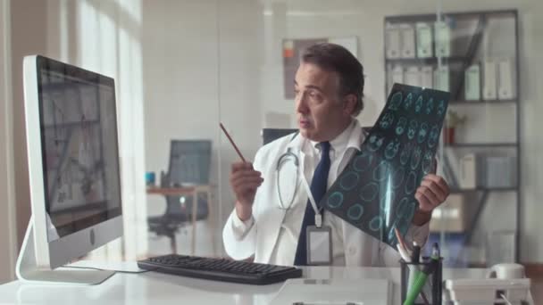 診療所でコンピュータを使用してビデオで呼びながら同僚にCtを示す高齢のX線技術者の中型ショット — ストック動画