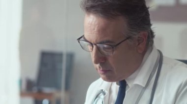 Klinikteki bilgisayarı kullanarak hastanın tıbbi kartını dolduran ciddi ve olgun bir pratisyen hekimin görüntüsünü aşağı eğ.