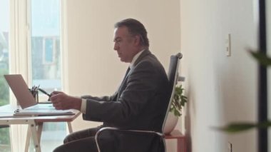 Ofis masasında kablosuz bilgisayarın önünde oturan takım elbiseli kıdemli çalışanın yan görüntüsü.