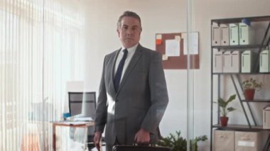 Kafkas CEO 'sunun evrak çantasını tutarken ve ofisteki kameraya bakarken orta uzunlukta portre fotoğrafı.