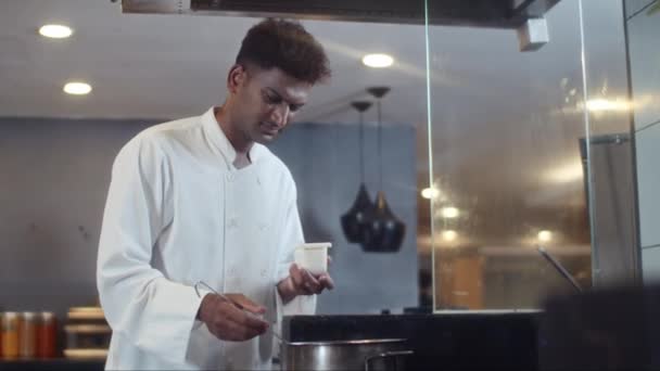 ホテルのキッチンで調理しながらスープに塩を加える白い制服を着たインドのステーションシェフの中型ショット — ストック動画