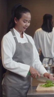 Önlük giymiş, sebze doğrayan ve restoran mutfağında kameraya bakan Asyalı bir kadının orta boy portresi.
