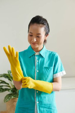 Asyalı genç temizlikçi üniformasını giyiyor ve lastik eldivenleri giymeye hazır.