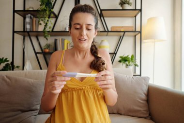 Oturma odasındaki kanepede oturan ve hamilelik testine pozitif sonuç alan mutlu genç bir kadın.