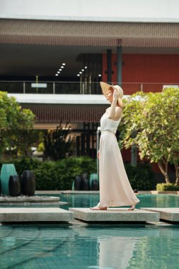 Genç beyaz bir kadının güneşli bir yaz gününün tadını çıkarırken otelin bölgesinde havuz başında yürüdüğünü gösteren dikey bir görüntü.