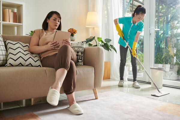 Азиатская женщина отдыхает на диване с книгой в то время как профессиональный уборщик от уборки делает работу по дому