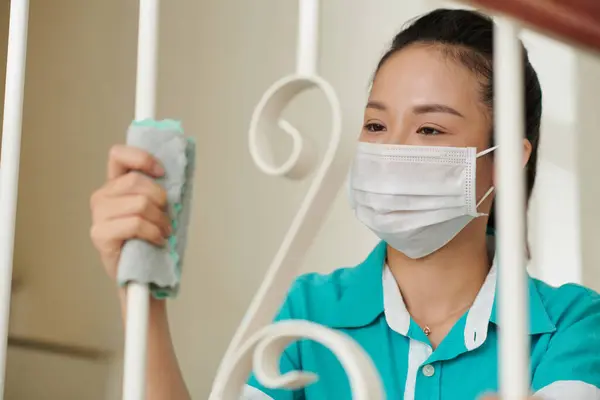 Asian young cleaner in mask wiping railings with rag during wet cleaning at home