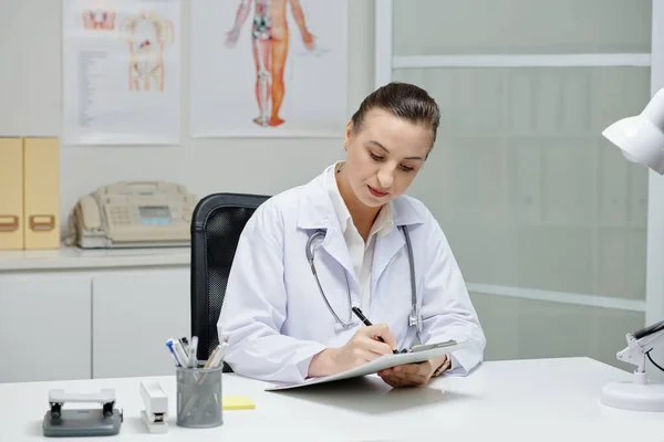 ホワイトコートの若い女性医師は オフィスの職場に座ってクリップボードに処方箋を書いています — ストック写真