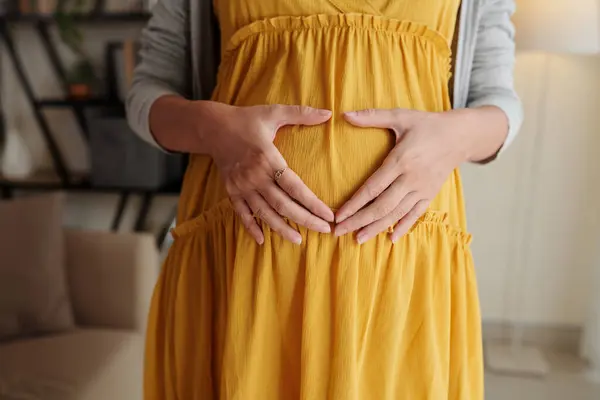 黄色いドレスの若い妊婦のクローズアップは 彼女の腹の上に手で心の形を作る 彼女は愛と赤ちゃんを期待しています — ストック写真