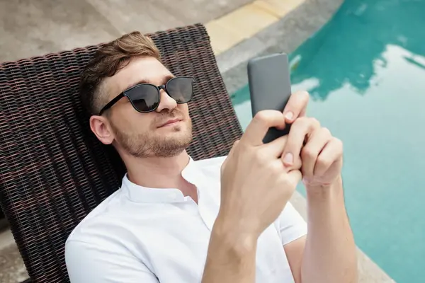 Handsome young adult Caucasian man wearing sunglasses relaxing on deck chair at poolside scrolling news feed in social network on smartphone