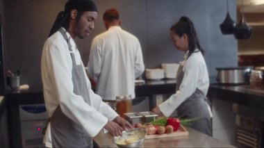 Kâsede sos hazırlayan ve restoran mutfağında iş arkadaşlarıyla birlikte kameraya bakan siyah aşçının orta boy portresi.
