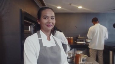 Restoranın mutfağında kameraya bakan önlük giymiş neşeli Asyalı kadın yardımcı şefin orta boy portresi.