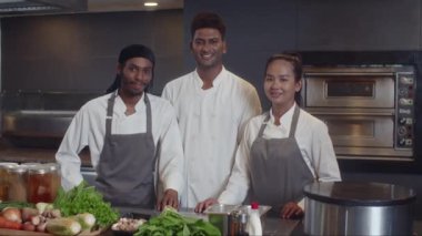 Mutfak personelinin orta boy portresi. Sebzelerle masanın yanında durup restoranda kameraya bakıyorlar.