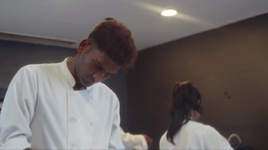 Beyaz üniformalı dalgın aşçı yamağının yemek pişirirken soğan doğrarken iş arkadaşlarıyla ağır çekimde...