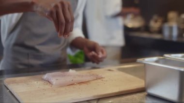 Beyaz üniformalı ve gri önlüklü dalgın aşçı yardımcısının restoranda yemek pişirirken balığa biber eklediği el kamerası görüntüsü.