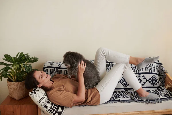ソファーに横たわっているときにかわいい小さな犬と遊ぶ陽気な若い女性 — ストック写真