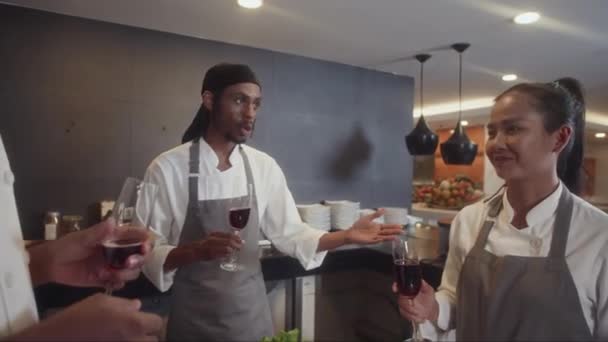 レストランキッチンでお祝いしながら赤ワインを飲む陽気なグルメの専門家のチームのハンドヘルドショット — ストック動画