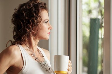 Düşünceli, olgun bir kadın bir fincan kahve içip pencereden dışarı bakıyor.