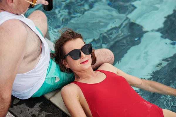 Smiling mature woman in red swimsuit and sunglasses resting head on laps of her boyfriend