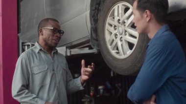 Orta uzunluktaki Afrikalı Amerikan lastik tamircisi araba servisi istasyonundaki adama tekerlek tamirini açıklıyor.