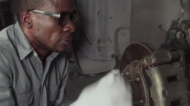 Araba servisinde araba tamir ederken gri üniformalı, vida gevşeten siyah erkek tamircinin el kamerası görüntüsü.