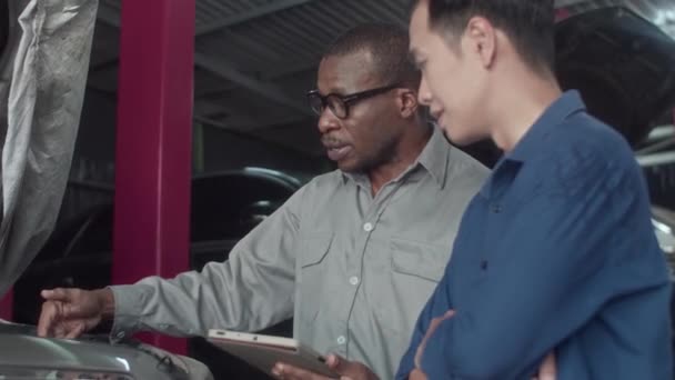 ガレージで車のエンジンを調べながら デジタルタブレットを見ているクライアントと修理担当者の中型ショット — ストック動画