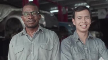 Gri üniformalı Afro-Amerikan ve Asyalı tamircilerin portresi araba servis istasyonunda kameraya gülümsüyor.