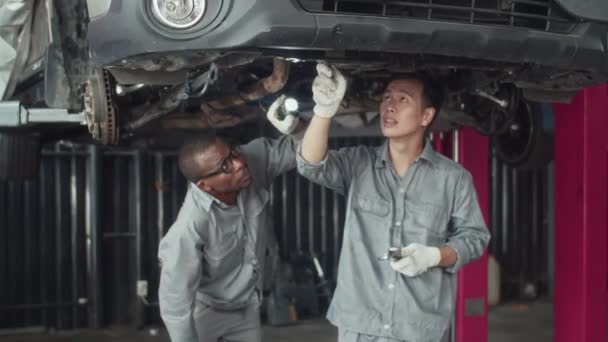 两个身着灰色制服 手电筒检查车底的多元文化机械师的中长镜头 — 图库视频影像