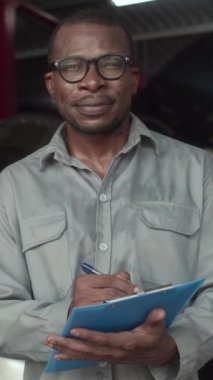 Neşeli siyah servis uzmanının dikey portresi araba tamiri ve kameraya bakmak üzerine doküman dolduruyor.