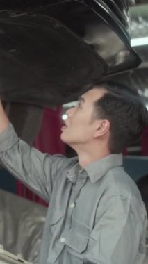 Dijital tablet kullanarak arabanın altını incelerken gri üniformalı Asyalı bir erkeğin dikey görüntüsü.