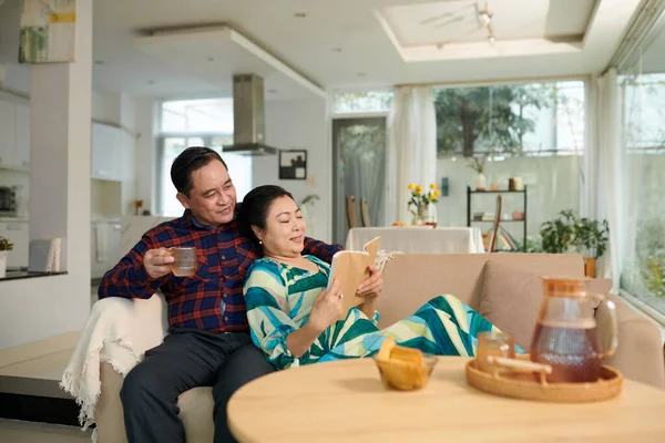 Senior Asian couple reading book and drinking tea when spending weekend at home