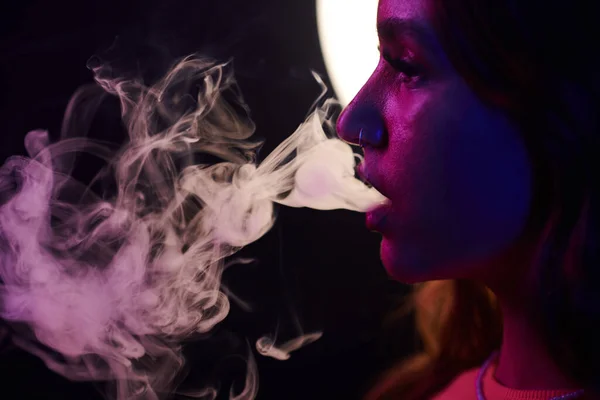 Young woman in neon light exhaling smoke of electronic cigarette