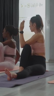 Dikey çekimde Afrikalı Amerikalı ve beyaz kadınlar fitness stüdyosunda omurga bükme egzersizi yapıyorlar.
