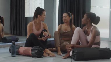 Yoga dersinden sonra stüdyoda yerde otururken birbirleriyle iletişim kuran üç çok kültürlü sporcu kadın.