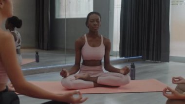 Orta uzunluktaki Afrikalı Amerikalı sporcunun Lotus pozisyonunda dinlenirken stüdyoda grup sınıfında yoga yaparken.