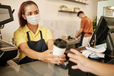 Kahveci barista, müşteriye kahve satarken tıbbi maske takıyor.