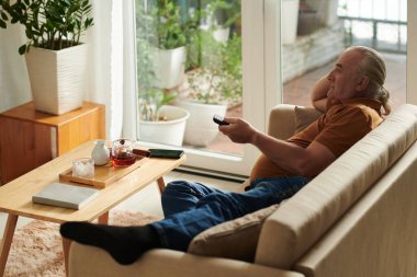 Yaşlı bir adam evdeki kanepede dinleniyor ve televizyonda yeni bir dizi ya da film izliyor.
