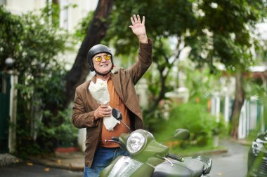 Mobiletinde duran ve kız arkadaşına el sallayan bir buket çiçekle mutlu bir son sınıf öğrencisi.