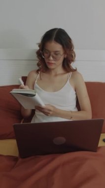 Dişi zoomer 'ın evde ders çalışırken dizüstü bilgisayarla yatakta oturduğu dikey görüntüsü.