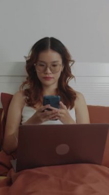 Genç bir kızın laptopun önünde yatakta çalışırken telefonla mesaj atarken dikey görüntüsü.