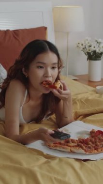 Asya kökenli genç bir kadının evde yatağında dinlenirken pizza yerken çekilmiş dikey bir fotoğrafı.