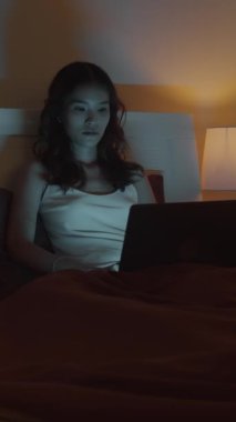 Kadın Asyalı işkoliğin kablosuz bilgisayar kullanarak gece yatakta rapor verirken dikey görüntüsü.