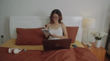 Bayan öğrencinin evde ders çalışırken dizüstü bilgisayarın başında oturup not aldığı orta boy bir fotoğraf.