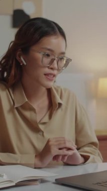 Asyalı genç bir kadının çevrimiçi toplantı sırasında iş arkadaşlarıyla konuşurken, evde dizüstü bilgisayarın önünde otururken dikey görüntüsü.